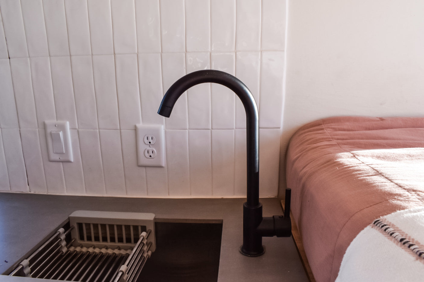 RV rental kitchenette features black faucet, white tiled backsplash, grey counter, and pink bed.
