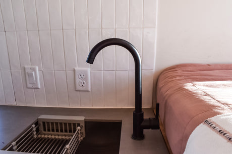 RV rental kitchenette features black faucet, white tiled backsplash, grey counter, and pink bed.