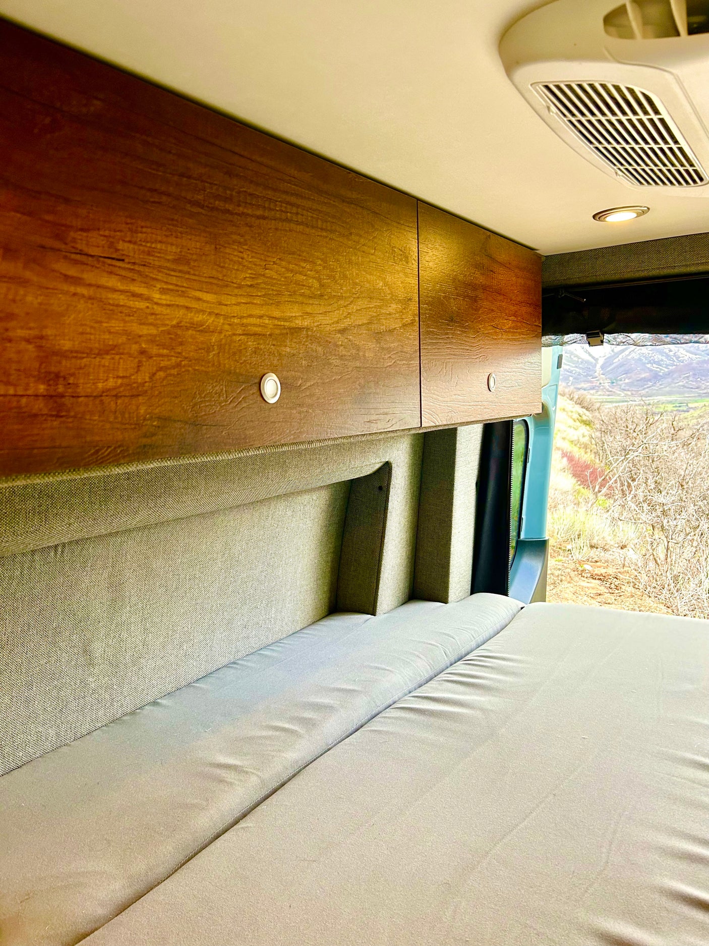 Inside an RV rental: light gray bed cushions, wooden cabinets, ceiling vent, and a window with mountain views.