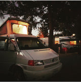 A white VW pop-top camper van with an illuminated top, parked at night in an RV park. Perfect for an RV rental adventure.