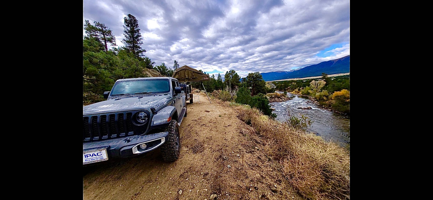 Grey Jeep Gladiator with rooftop tent parked by a river, mountains in distance. An RV rental alternative for adventure.
