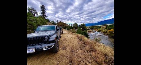 Grey Jeep Gladiator with rooftop tent parked by a river, mountains in distance. An RV rental alternative for adventure.