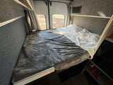 RV rental: comfortable bed in a camper van interior with grey and white bedding. Rear windows show an outside view.