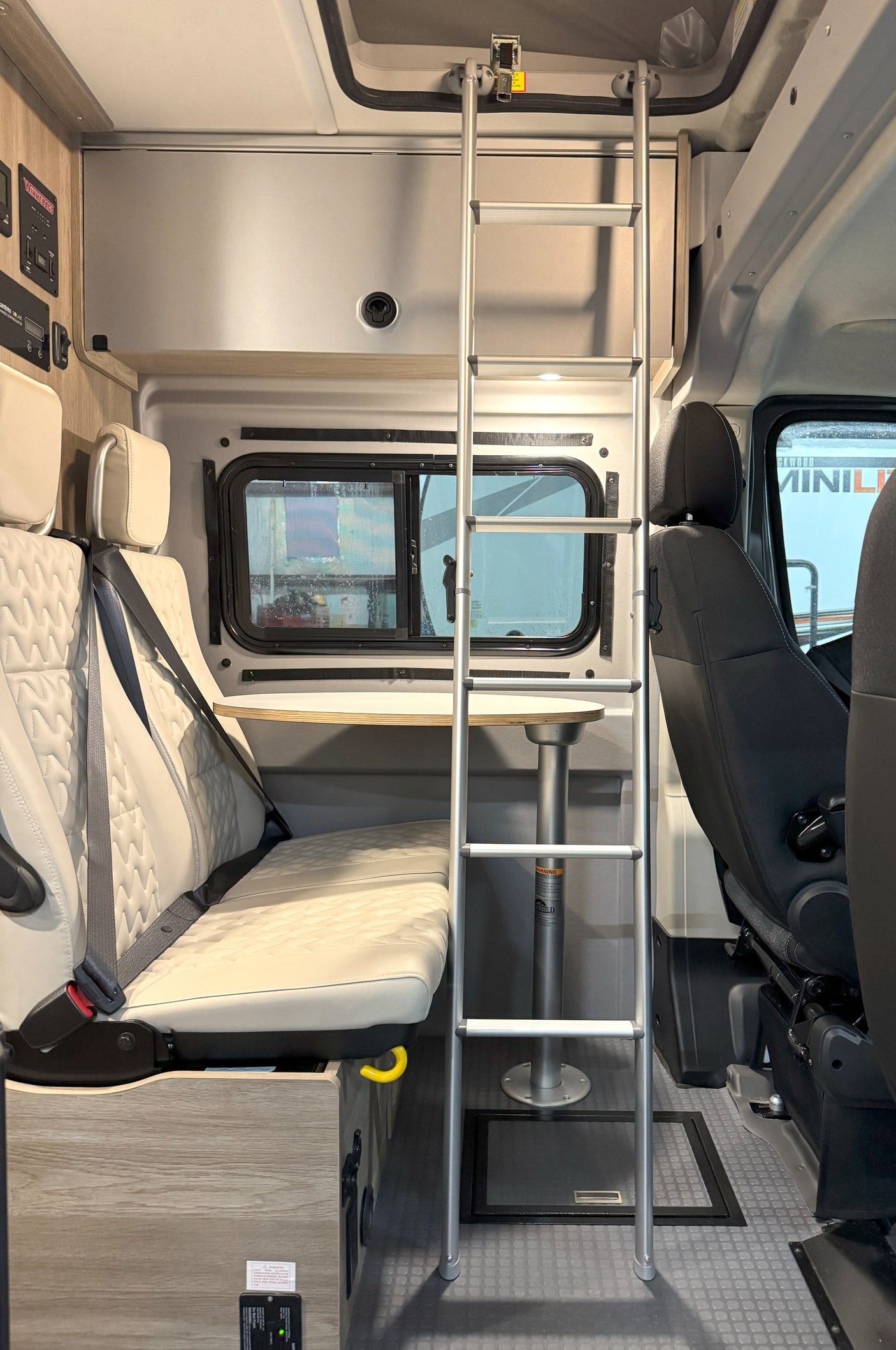 Inside an RV rental van: seating with seatbelts, a detachable table, a window, and a ladder to an upper sleeping area.