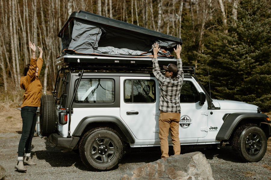 Two people set up a rooftop tent on a white RV rental Jeep Wrangler for an outdoor camping adventure.