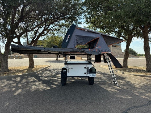 Ready to camp RV rental: White off-road trailer with black rooftop tent & awning deployed in a parking lot.