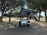 Ready to camp RV rental: White off-road trailer with black rooftop tent & awning deployed in a parking lot.