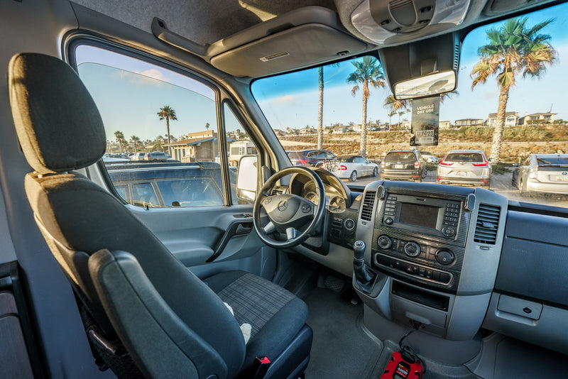 Inside an RV rental van showing the driver's seat and dashboard. Outside, a coastal parking lot with palm trees and a day-use pass hangs. — photo 9