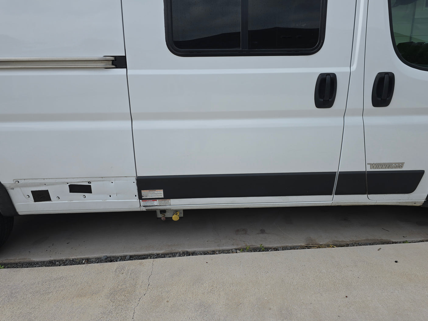 Side view of a white Winnebago RV rental, showing sliding door, windows, and external utility connections.