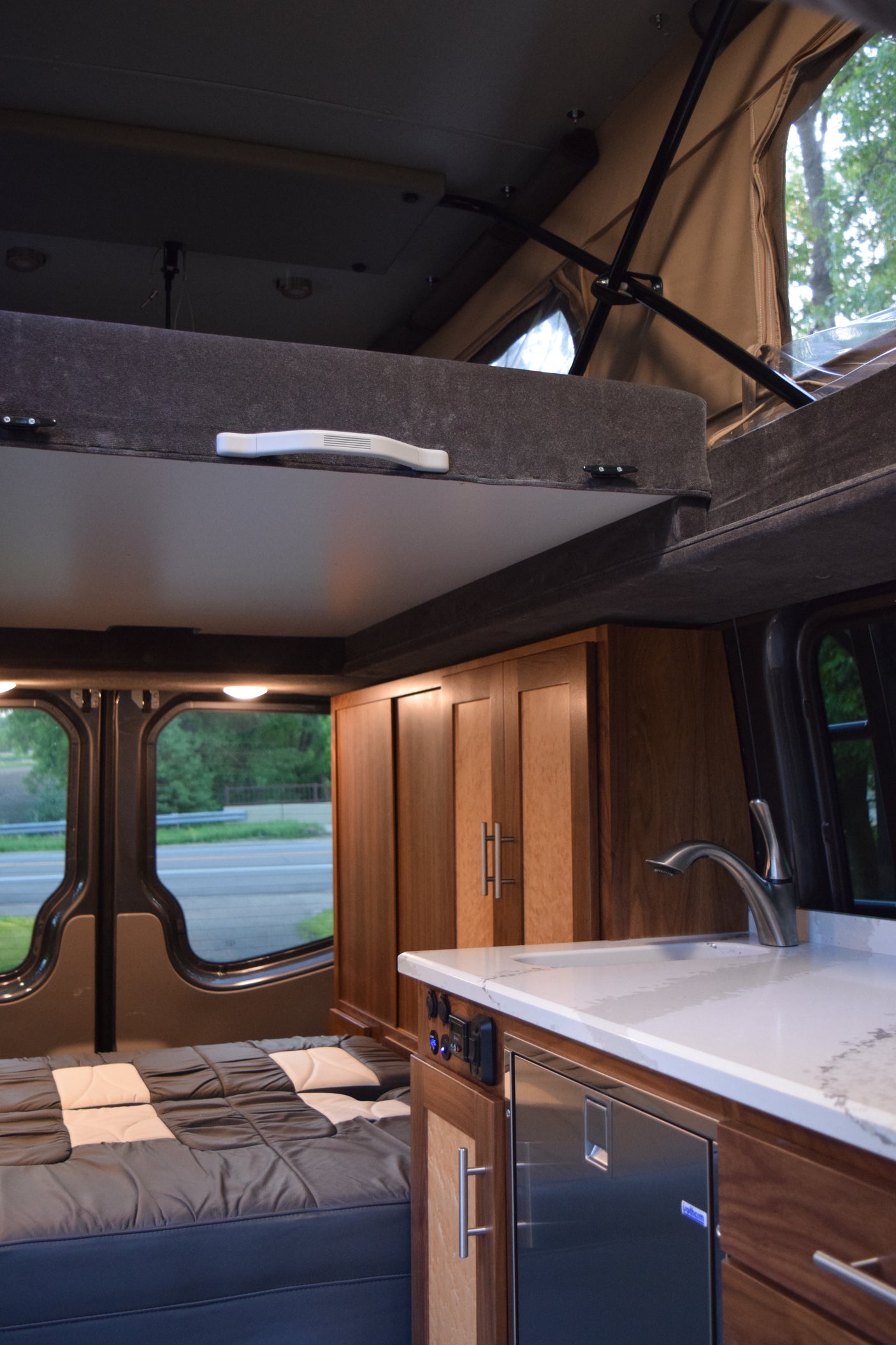 An RV rental interior featuring a comfy bed, modern kitchen with sink, refrigerator, and wood cabinets, plus a pop-top.