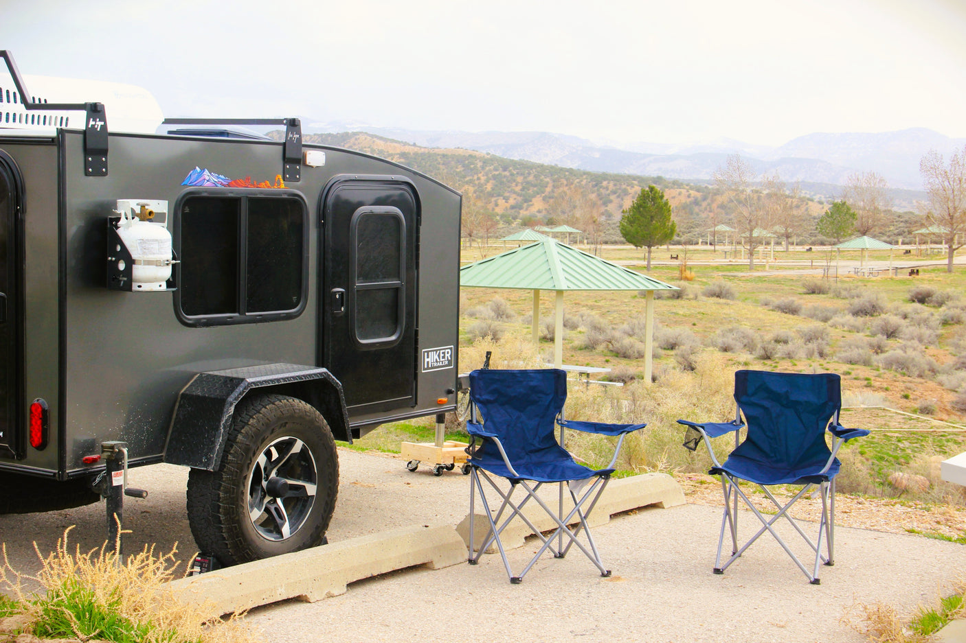 Explore nature with this compact RV rental. Teardrop trailer and camping chairs at a scenic park campsite.