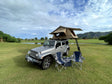  mountains in Hawaii background. Ideal for an RV rental adventure.|Adventure-ready silver Jeep with TMBK rooftop tent & camp chairs on grassy field. Explore with our RV rental!|An ideal beach camp setup: two chairs