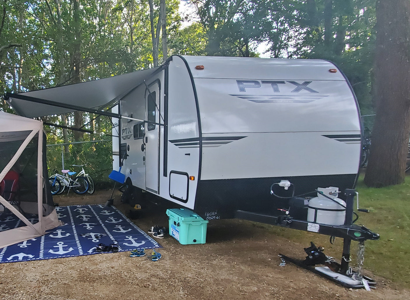 This white RV rental is set up with an extended awning and screen tent at a sunny, wooded campsite, ready for camping.
