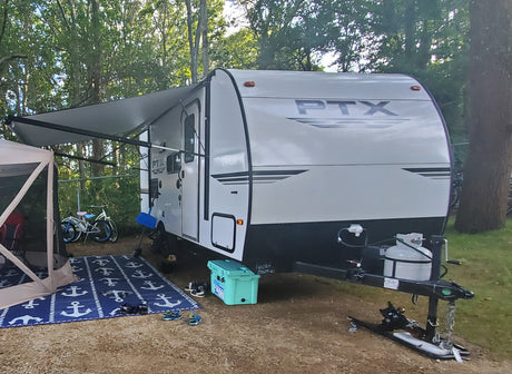 This white RV rental is set up with an extended awning and screen tent at a sunny, wooded campsite, ready for camping.