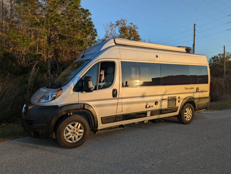 A silver Winnebago Tellaro rv rental van with a small dog in the driver's seat, parked on an asphalt road at sunset.