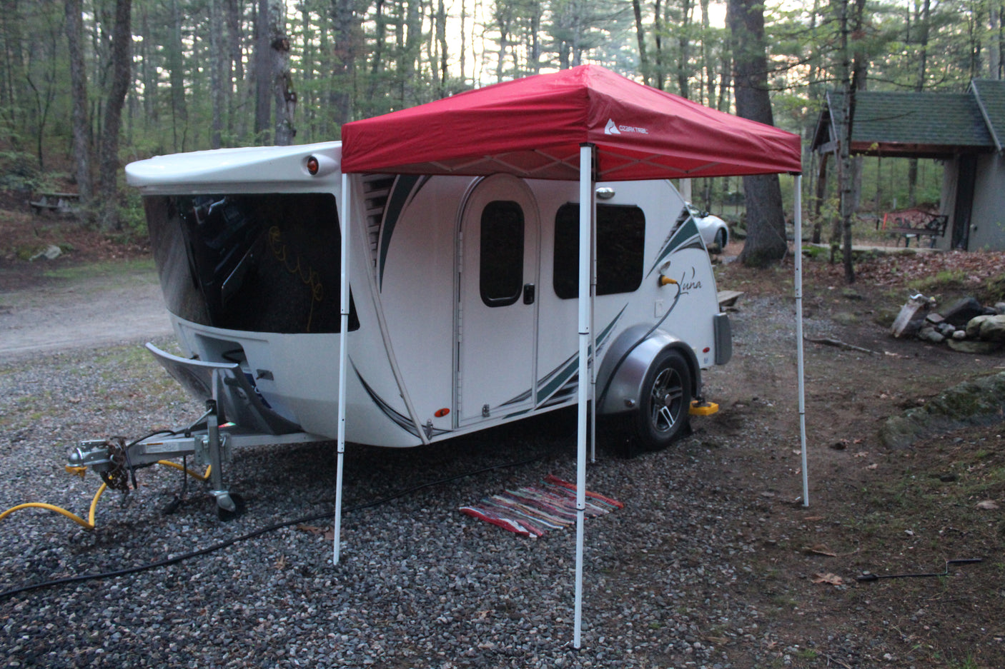 This RV rental shows a white teardrop camper under a red canopy, set up for camping in a rustic, wooded spot.