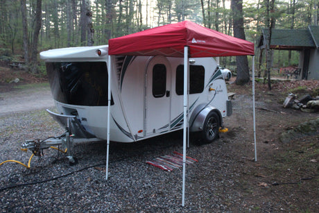 This RV rental shows a white teardrop camper under a red canopy, set up for camping in a rustic, wooded spot.