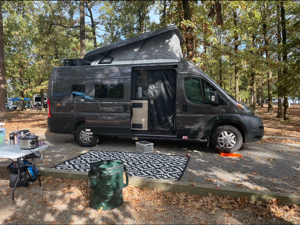 Spacious dark gray rv rental camper van with pop-top set up at a leafy campground. Perfect for outdoor adventure.|Dark gray pop-top rv rental camper van parked at a gravel campsite with a blue canopy & outdoor kitchen.|Dark gray Winnebago Solis RV rental parked on grass with doors open and awning out