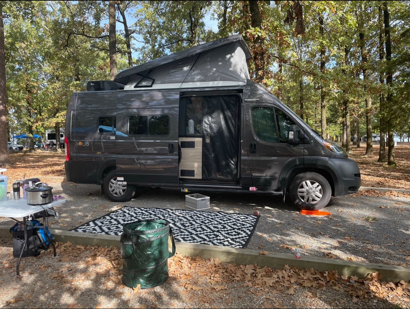 Spacious dark gray rv rental camper van with pop-top set up at a leafy campground. Perfect for outdoor adventure.|Dark gray pop-top rv rental camper van parked at a gravel campsite with a blue canopy & outdoor kitchen.|Dark gray Winnebago Solis RV rental parked on grass with doors open and awning out — photo 1