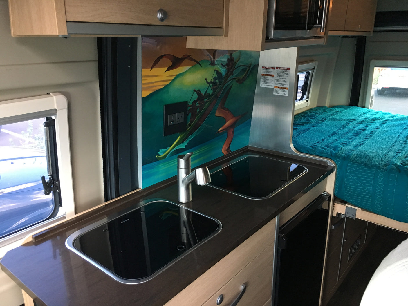 An RV rental's clever interior: compact kitchenette with two sinks/cooktops, vibrant mural, and a comfy bed for travelers.