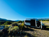 A silver camper van (RV rental) is parked with open doors in a scenic, mountainous landscape under a blue sky.