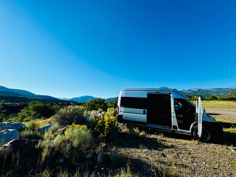 A silver camper van (RV rental) is parked with open doors in a scenic, mountainous landscape under a blue sky.