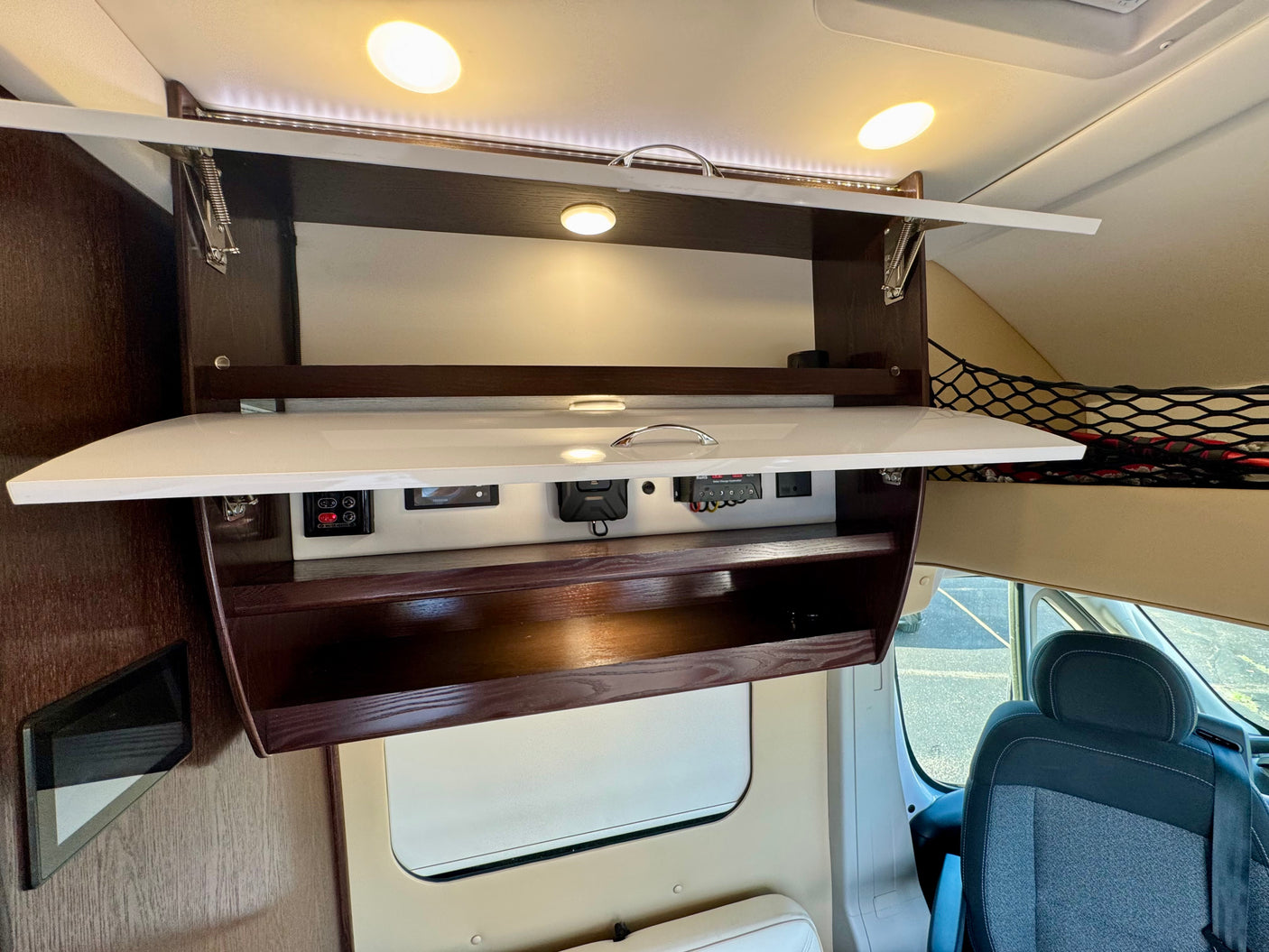 View inside an rv rental, featuring an open overhead cabinet with integrated controls, bright lighting, and visible passenger seating.