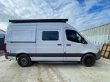 This white RV rental camper van features a rooftop awning, side step, and tinted windows, parked on concrete under a cloudy sky.
