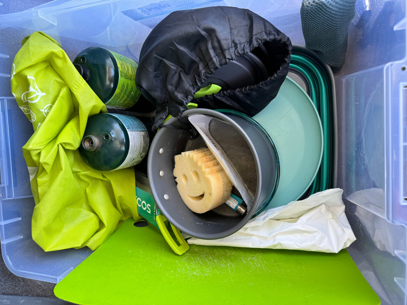Packed RV rental camping supplies: propane tanks, cookware, collapsible dishes, and a smiley sponge in a clear tote.