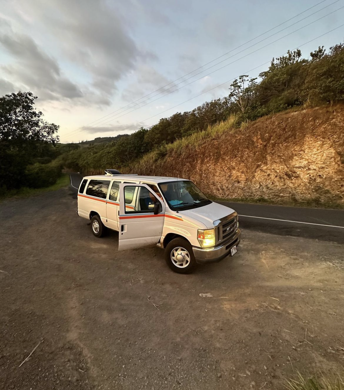 Experience van life! A white RV rental van, door open, parked on a dirt shoulder in a lush, hilly natural setting.