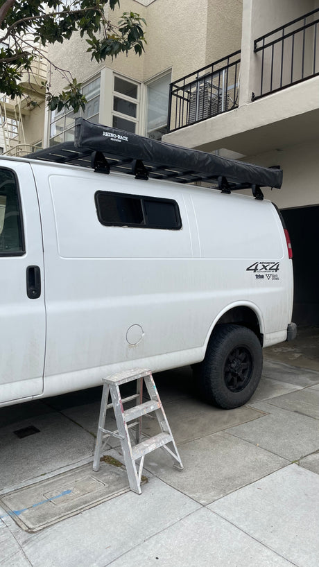 White 4x4 camper van (potential RV rental) with roof rack awning, parked. A metallic step ladder stands nearby.