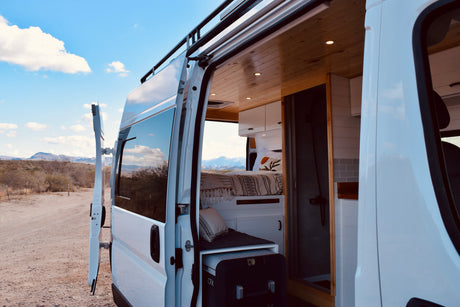 Open door of an RV rental campervan, revealing a cozy, custom interior with a bed and wooden ceiling, set in a desert landscape.