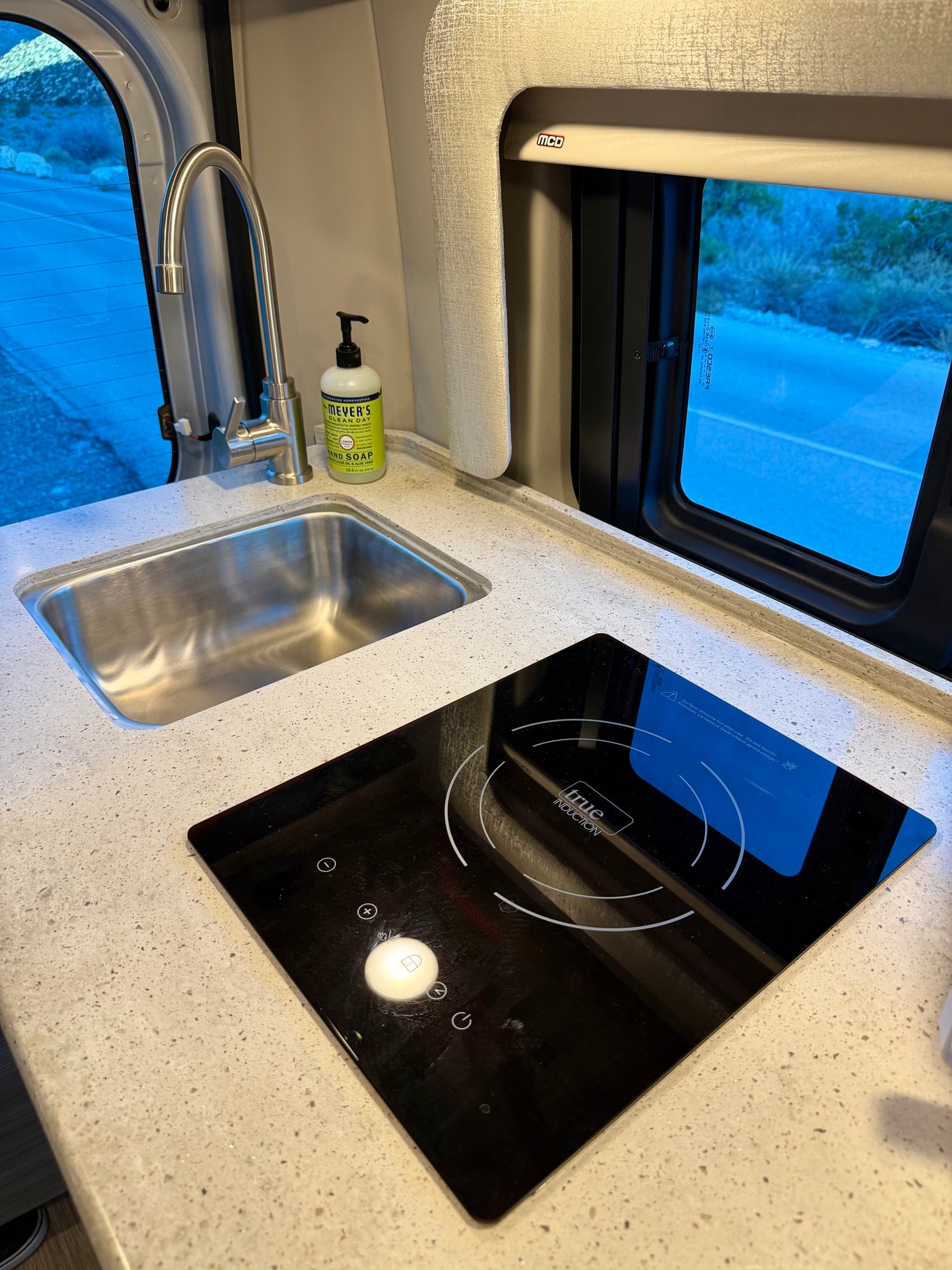RV rental kitchen: a sleek sink, induction cooktop, Mrs. Meyer's soap, and window revealing a road and nature.