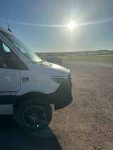 White Sprinter rv rental van on gravel road with green fields and hills under bright sun.
