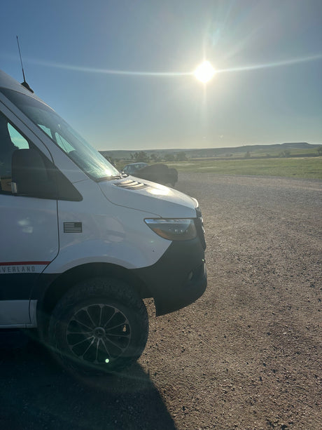 White Sprinter rv rental van on gravel road with green fields and hills under bright sun.