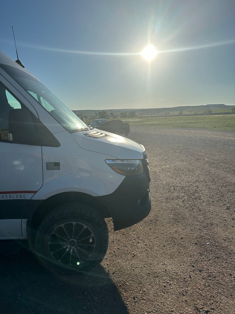  sits centered on a dirt path in a sunny pine forest campsite with large rocks.|A customized Mercedes-Benz Sprinter van — photo 7