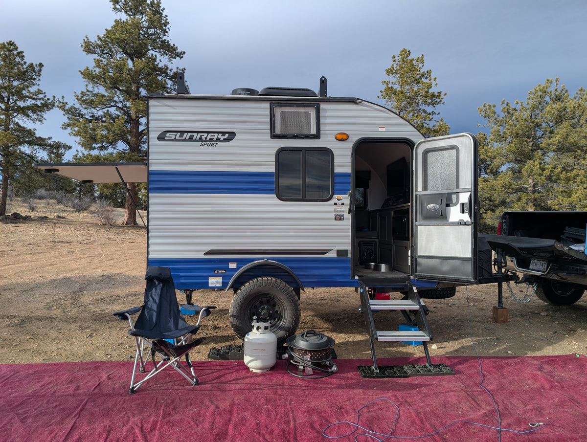 Sunray Sport rv rental travel trailer with awning extended, door open, and outdoor living area on a red rug at a wooded campsite. — photo 2