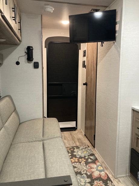 An RV rental interior with a light-colored sofa, wall-mounted TV, floral rug, and exit door. A compact living space.