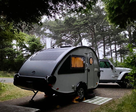 Teardrop RV rental, equipped with solar panels, parked with a white Jeep Wrangler in a serene, misty forest campsite.