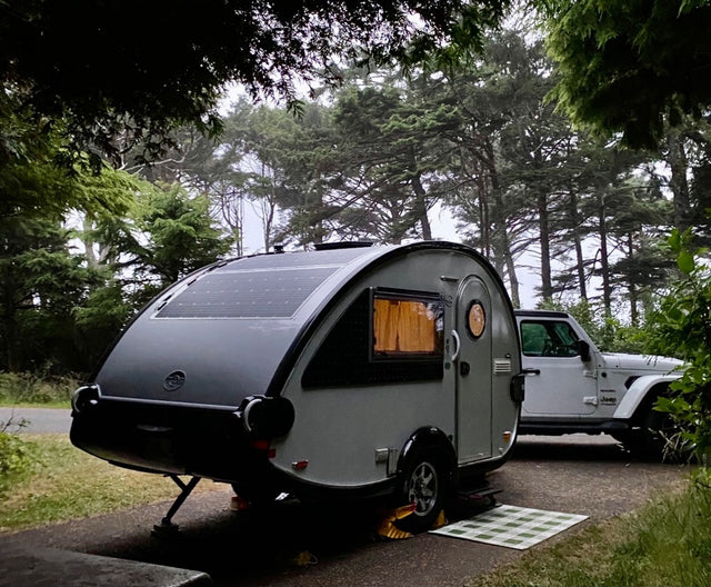 Teardrop RV rental, equipped with solar panels, parked with a white Jeep Wrangler in a serene, misty forest campsite.
