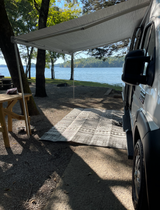 Relaxing RV rental setup by the lake with an extended awning, mat, and picnic table at a scenic campsite.