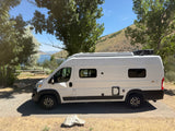 A white Winnebago Solis camper van, an ideal RV rental, parked by a scenic lake and hills. Perfect for travel adventures.