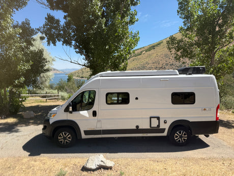 A white Winnebago Solis camper van, an ideal RV rental, parked by a scenic lake and hills. Perfect for travel adventures.