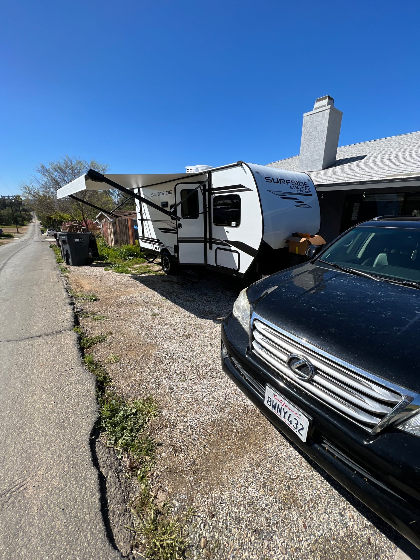 Sunny view: a white Surfside RV, possibly an rv rental, parked on a gravel driveway beside a house, with a black car in the foreground.