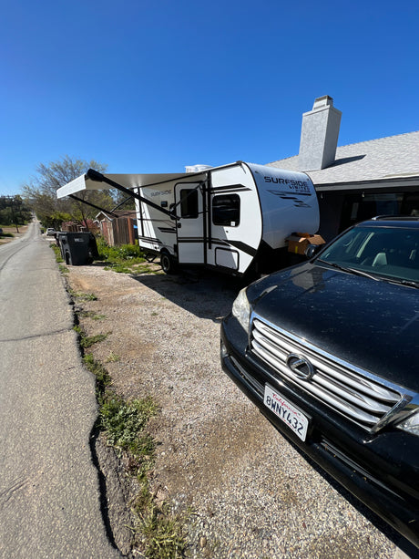 Sunny view: a white Surfside RV, possibly an rv rental, parked on a gravel driveway beside a house, with a black car in the foreground.