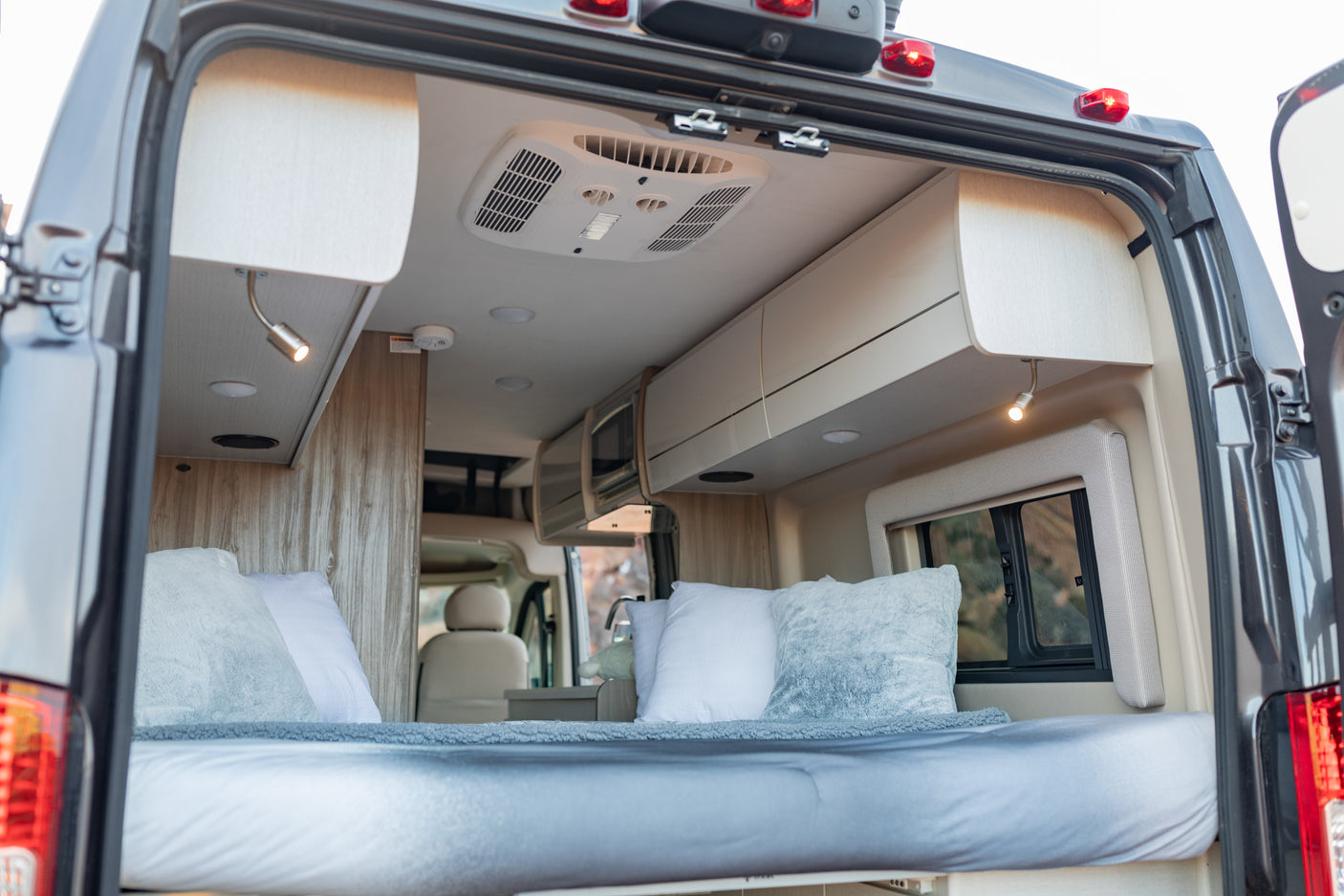 Inside an RV rental: a modern, light-filled sleeping area with a bed, overhead storage, and climate control, seen from open rear doors.