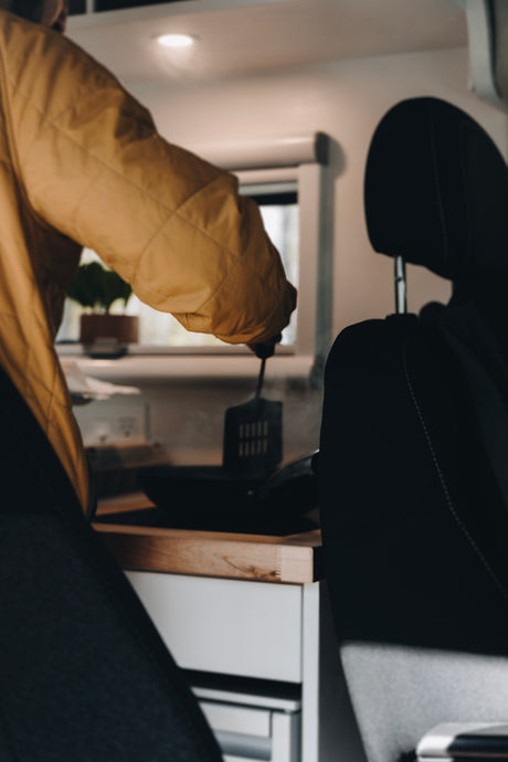 Person cooking in an RV rental kitchen, stirring food in a steaming pan. A vehicle seat is in the foreground.