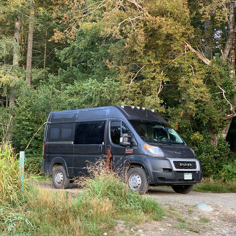Dark grey RV rental camper van, a Ram ProMaster, parked on a gravel path near a dense forest.