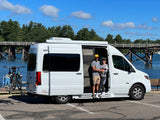 Happy couple with their dog in a white RV rental, parked by a lake with a wooden bridge and bikes.