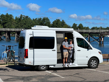 Happy couple with their dog in a white RV rental, parked by a lake with a wooden bridge and bikes.